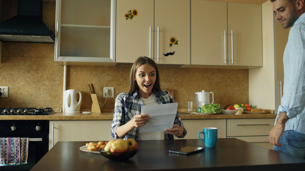 Une femme lit un courrier dans sa cuisine et sourit, surprise